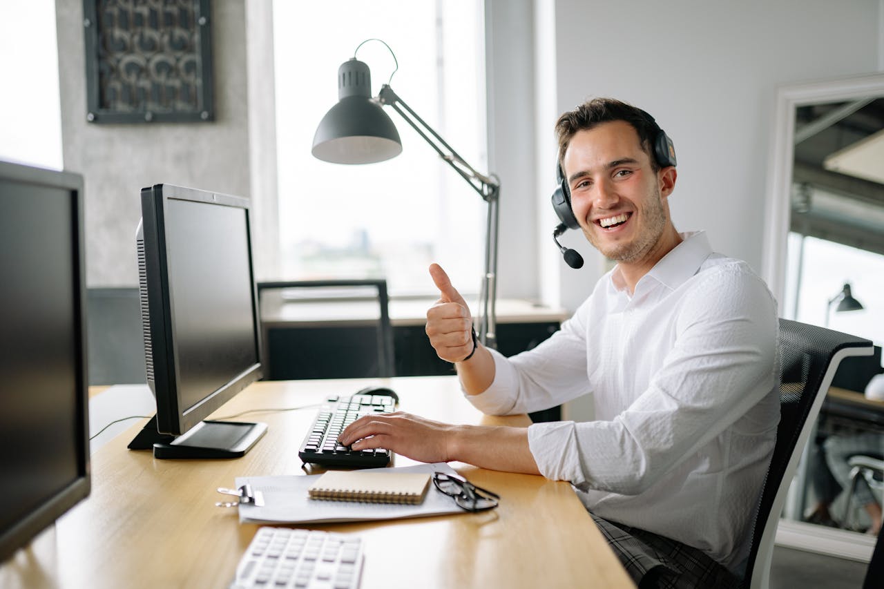 hero-img-02 Happy man in office wearing headset, giving a thumbs up while typing at a computer.